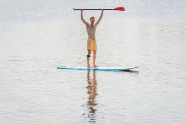 full length of overjoyed redhead man in yellow swim shorts holding paddle in raised hands while standing on sup board on lake with calm water on summer weekend day clipart
