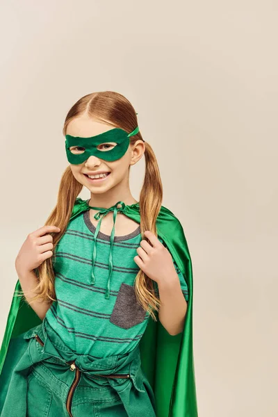 笑顔の女の子です緑のスーパーヒーローの衣装でマントとマスク顔に 双子の尾の髪型で彼女の髪に触れるとグレーの背景に国際子供の日を祝います — ストック写真