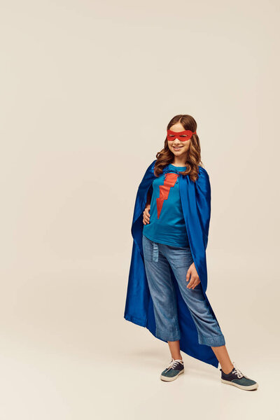 счастливая девушка в костюме супергероя в синем плаще и красной маске, стоящая с рукой на бедре и позируя в джинсах и футболке во время празднования Дня защиты детей на сером фоне 