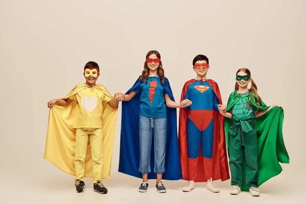 happy interracial preteen kids in colorful superhero costumes with cloaks and masks holding hands and looking at camera while celebrating Child protection day holiday on grey background in studio