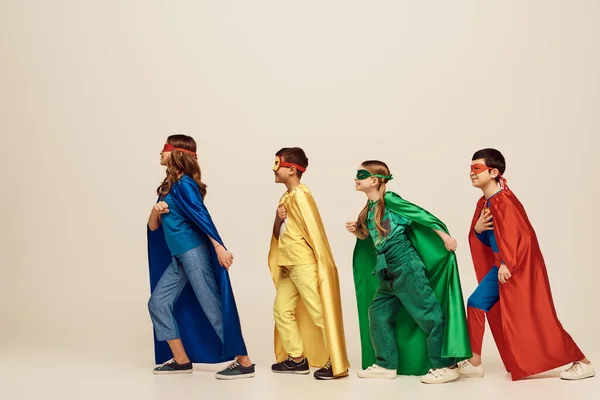 side view of happy interracial kids in colorful costumes with cloaks and masks smiling and walking together on grey background in studio, Child Protection Day concept
