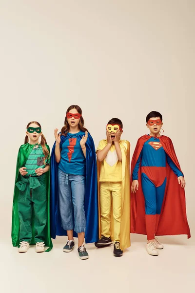 shocked interracial children in colorful superhero costumes with cloaks and masks looking at camera on grey background in studio, Child Protection Day concept