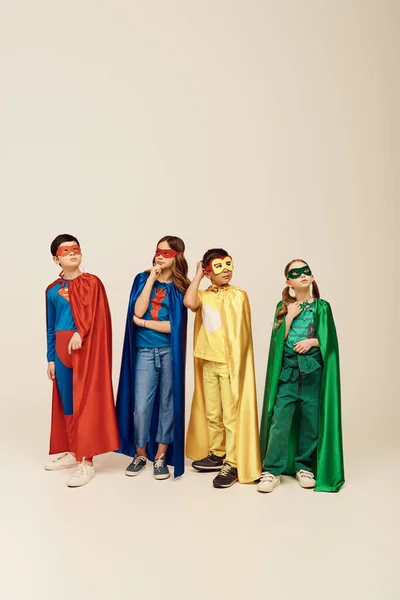 pensive multicultural kids in colorful superhero costumes with cloaks and masks scratching head and looking away while thinking on grey background in studio, children\'s day concept