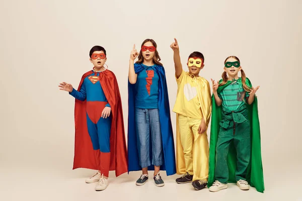 multicultural kids in colorful superhero costumes with cloaks and masks showing idea gesture with fingers and standing with opened mouth on grey background in studio, children\'s day concept