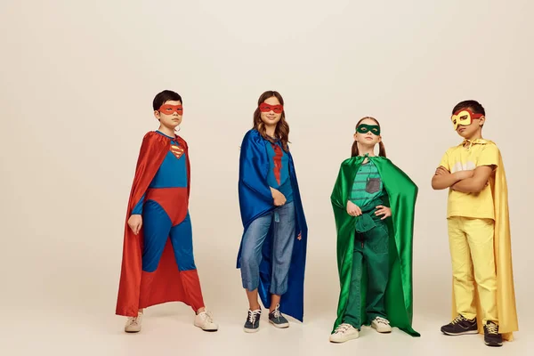 interracial kids in colorful superhero costumes with masks and cloaks standing and posing together on grey background in studio, International children\'s day concept