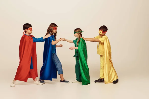 side view of interracial kids in colorful superhero costumes with masks and cloaks fighting with each other on grey background in studio, International children\'s day concept