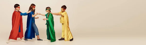 side view of multicultural kids in colorful superhero costumes with masks and cloaks fighting with each other on grey background in studio, International children\'s day concept, banner