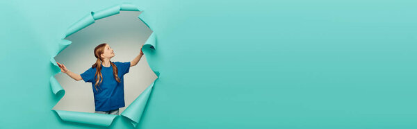 cheerful and redhead preteen girl in t-shirt smiling and looking at blue torn paper creating hole on white background, Happy children's day concept, banner 