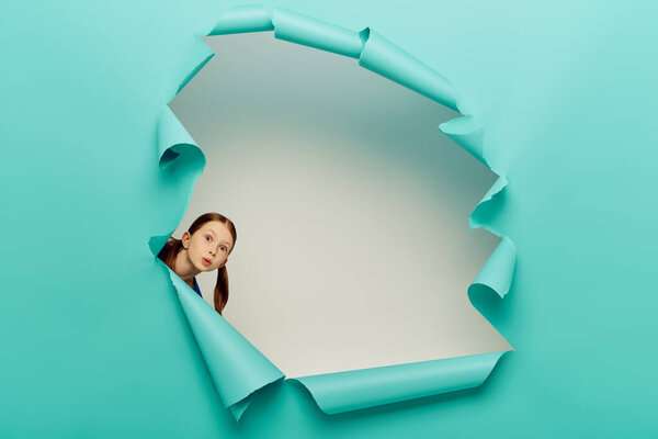 surprised redhead preteen girl looking at camera through blue torn paper hole on white background, International Child Protection Day concept 