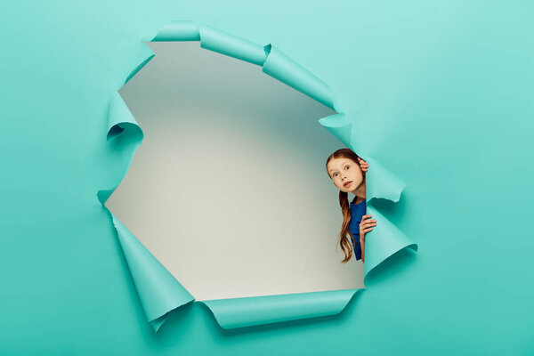 Испуганная рыжая девочка-подросток в голубой футболке смотрит в камеру, стоя за дырой в голубой бумаге на белом фоне