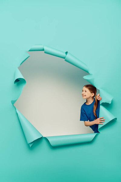 Веселая рыжеволосая девушка в футболке смотрит в сторону, прикасаясь к дыре в голубой газете и отмечая Международный день защиты детей на белом фоне