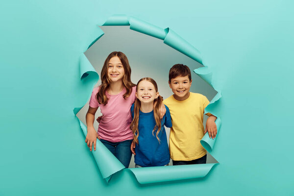 Positive interracial preteen kids in colorful t-shirts looking at camera while celebrating child protection day and standing behind hole in blue paper background