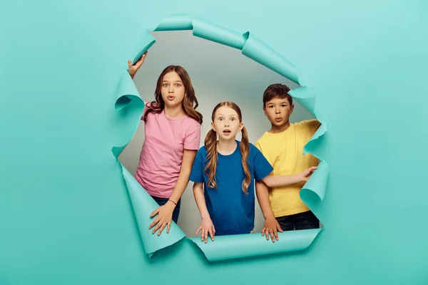 stock image Shocked multiethnic preteen kids in colorful t-shirts looking at camera while celebrating child protection day behind hole in blue paper background