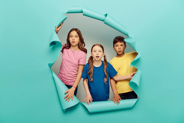 Shocked multiethnic preteen kids in colorful t-shirts looking at camera while celebrating child protection day behind hole in blue paper background