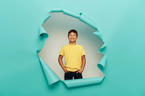 Smiling multicultural boy holding hands in pockets of pants and looking at camera while celebrating child protection day behind hole in blue paper background