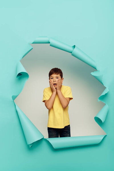 Shocked multiracial boy in yellow t-shirt touching cheeks and looking at camera during world child protection day while standing behind hole in blue paper background