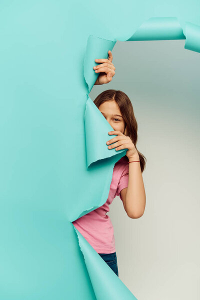 Preteen child in casual clothes covering face and looking at camera through hole in blue paper while hiding on grey background, International Child Protection Day concept 