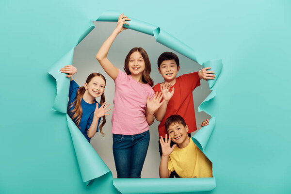 Smiling interracial kids in casual clothes waving hands at camera during international children day celebration behind hole in blue paper background