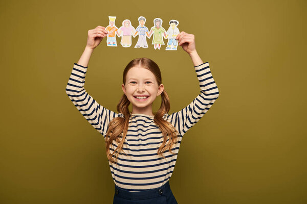 Smiling redhead girl in striped shirt holding drawing paper characters and looking at camera during child protection day celebration on khaki background