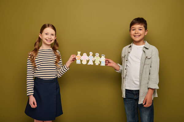 Smiling multiethnic preteen friends in casual clothes holding paper drawn characters and looking at camera during child protection day celebration on khaki background