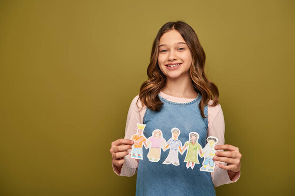 Smiling preteen girl in denim sundress holding drawn paper characters and looking at camera during child protection day celebration on khaki background