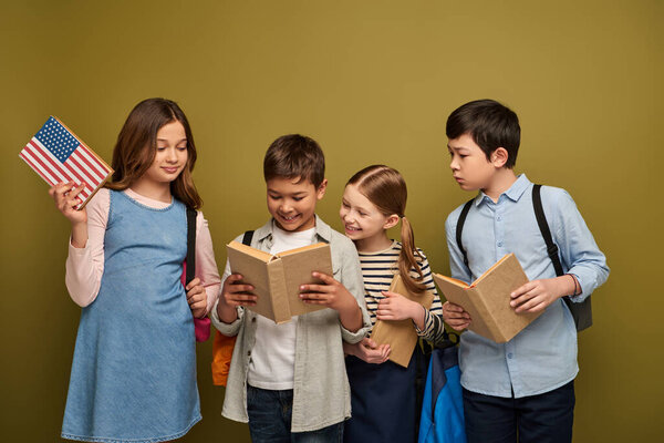Smiling multiethnic kids with backpacks looking at multiracial friend reading book, and girl holding dictionary with usa flag during child protection day celebration on khaki background