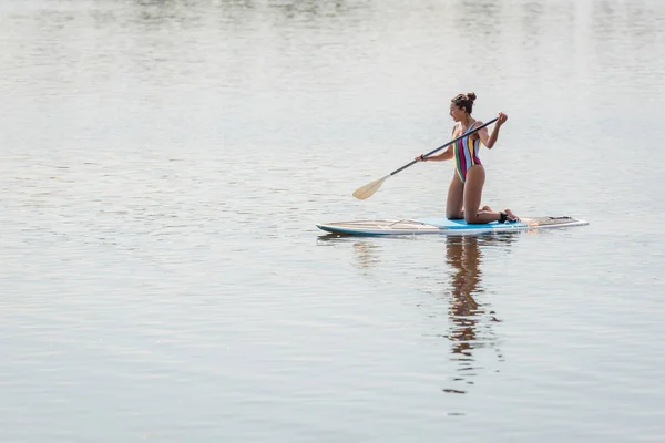 膝の上に立ち夏の日の週末に川の上でボード上でセーリングしながらカラフルな水着を保持パドルで細いアフリカ系アメリカ人女性 — ストック写真