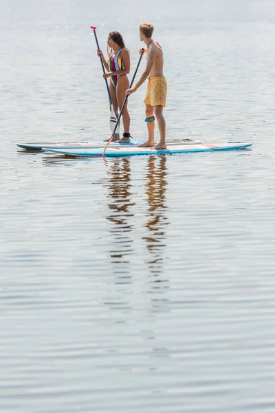 若いですRedhead男で泳ぐショートパンツとブルネットアフリカ系アメリカ人女性でカラフルな水着セーリングオンアップボード上の穏やかな川で夏の日 — ストック写真