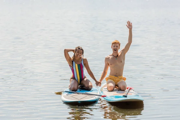 stock image overjoyed redhead man holding hands with smiling african american woman in striped swimsuit and showing hello gesture from sup boards on river in summer