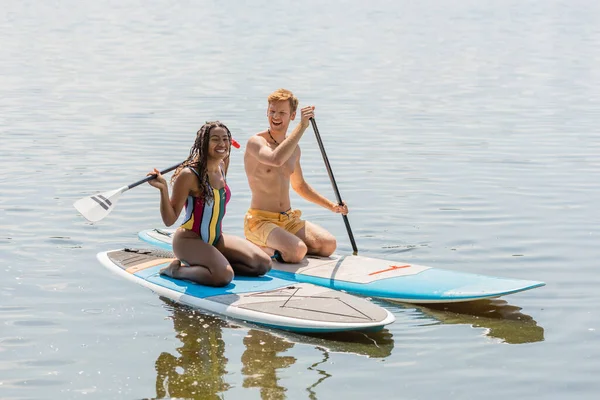 夏の週末に湖で過ごす間にボード上に座ってショートパンツを泳ぐ縞模様の水着と幸せな赤毛の男で喜びアフリカ系アメリカ人女性 — ストック写真