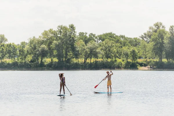 stock image full length of african american woman sailing on sup board and looking away near young and sportive man in swim shorts on scenic lake with green trees on shore