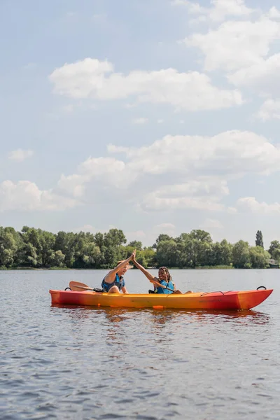 carefree interracial couple in life vests giving high five while spending summer weekend on river and sailing in sportive kayak along green riverside under blue sky with white clouds