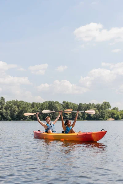 2人の陽気な異人種間の友人がパドルを持ち上げながら湖の絵のように青い空の下でカヤックに座って緑の川沿いを背景に — ストック写真
