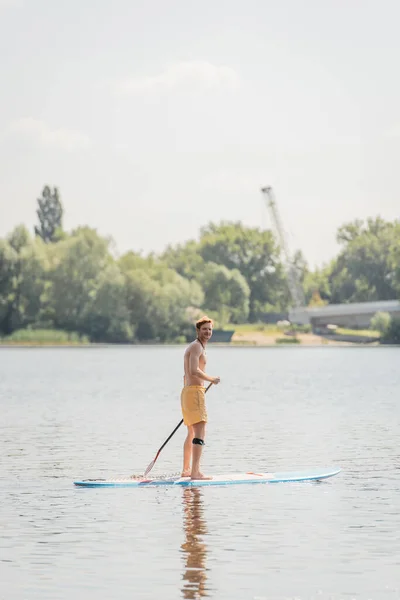 黄色のスポーツレッドヘッドの男の完全な長さはパドルを保持し 夏の週末の日に絵のように美しい湖を航海しながらカメラを見てショートパンツを泳ぐ — ストック写真