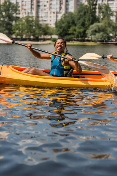 счастливая и спортивная африканская американка в спасательном жилете, держащая весло во время плавания на байдарке с юным другом во время летних выходных на озере в городе