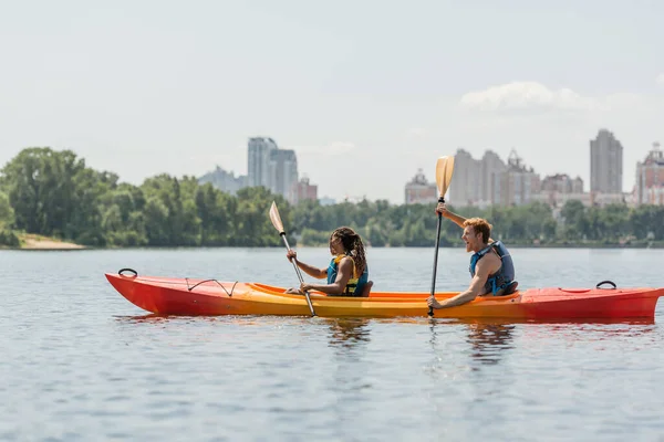 stock image side view of active african american woman and excited redhead man in life vests paddling in sportive kayak along riverside with picturesque cityscape on blurred background