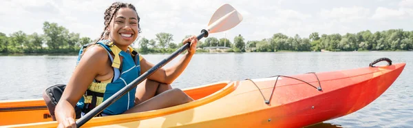 夏の週末を背景に緑の川沿いのカメラでパドルと笑顔でカヤックをしているアフリカ系アメリカ人女性 — ストック写真