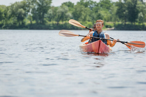 young redhead man in life vest holding paddle and looking away while sailing in sportive kayak with african american woman during summer weekend on river