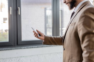 Çağdaş ofisteki büyük pencerelerin yanında boş ekranla cep telefonu tutan bej renkli ceketli sakallı işadamının kısmi görüntüsü