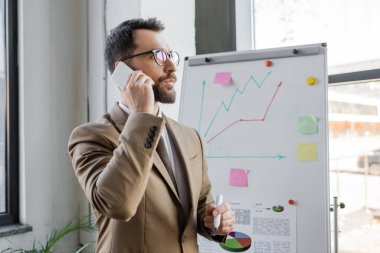 thoughtful manager in stylish business attire and eyeglasses standing with marker near analytics and graphs on flip chart and talking on mobile phone in office clipart