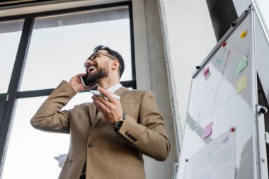 Düşük açılı, bej renkli spor ceketli iş adamının düşük açılı görüntüsü elinde keçeli kalemle akıllı telefondan bilgi grafikleri ve ofiste yapışkan notlarla konuşuyor.