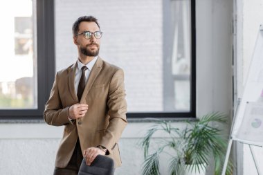 confident and bearded businessman in beige blazer, tie and eyeglasses standing near office chair, blurred flip chart and potted plant and looking away clipart