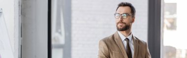 accomplished and confident bearded businessman in beige blazer, tie and stylish eyeglasses looking away while standing in contemporary office on blurred background, banner clipart