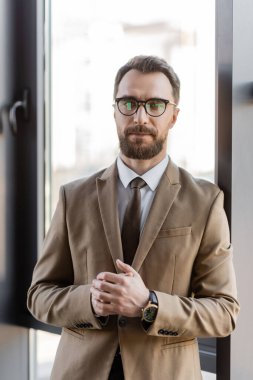 ambitious bearded businessman in beige blazer, stylish eyeglasses and luxury wristwatch looking at camera while standing with clenched hands near window in office clipart