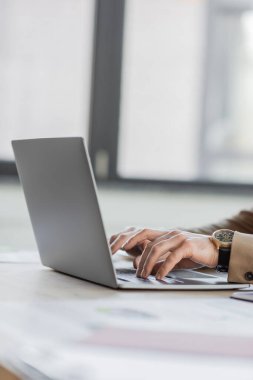 Bej rengi forma ve lüks kol saati giyen şık ve başarılı bir şirket yöneticisinin iş yerindeki laptopta yazı yazması, bulanık bir arka plan.