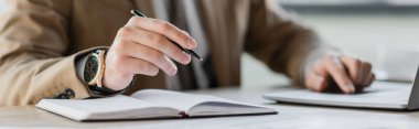 partial view of accomplished businessman in beige blazer and luxury wristwatch sitting with pen near notebook and laptop on work desk in office, banner clipart