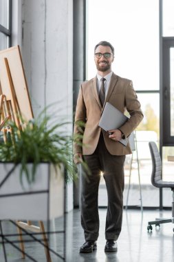 Moda ceket, pantolon, kravat ve gözlüklü başarılı sakallı işadamı dizüstü bilgisayar tutuyor ve modern ofisteki kamera önünde gülümsüyor.