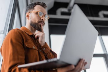 Moda gözlüklü ve tişörtlü ciddi sakallı işadamının düşük açılı görüntüsü dizüstü bilgisayara bakıyor ve ofiste bulanık bir şekilde duruyor. 