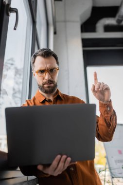 worried bearded businessman in stylish eyeglasses and shirt pointing up with finger while showing attention gesture during video conference on laptop in office, corporate lifestyle  clipart