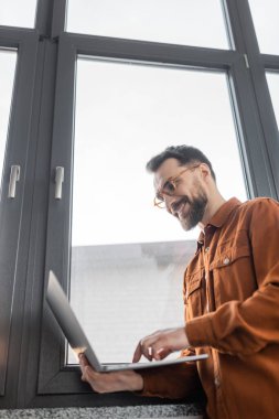 low angle view of successful and bearded businessman in eyeglasses and stylish shirt standing near window in modern office and smiling while using laptop, corporate lifestyle  clipart